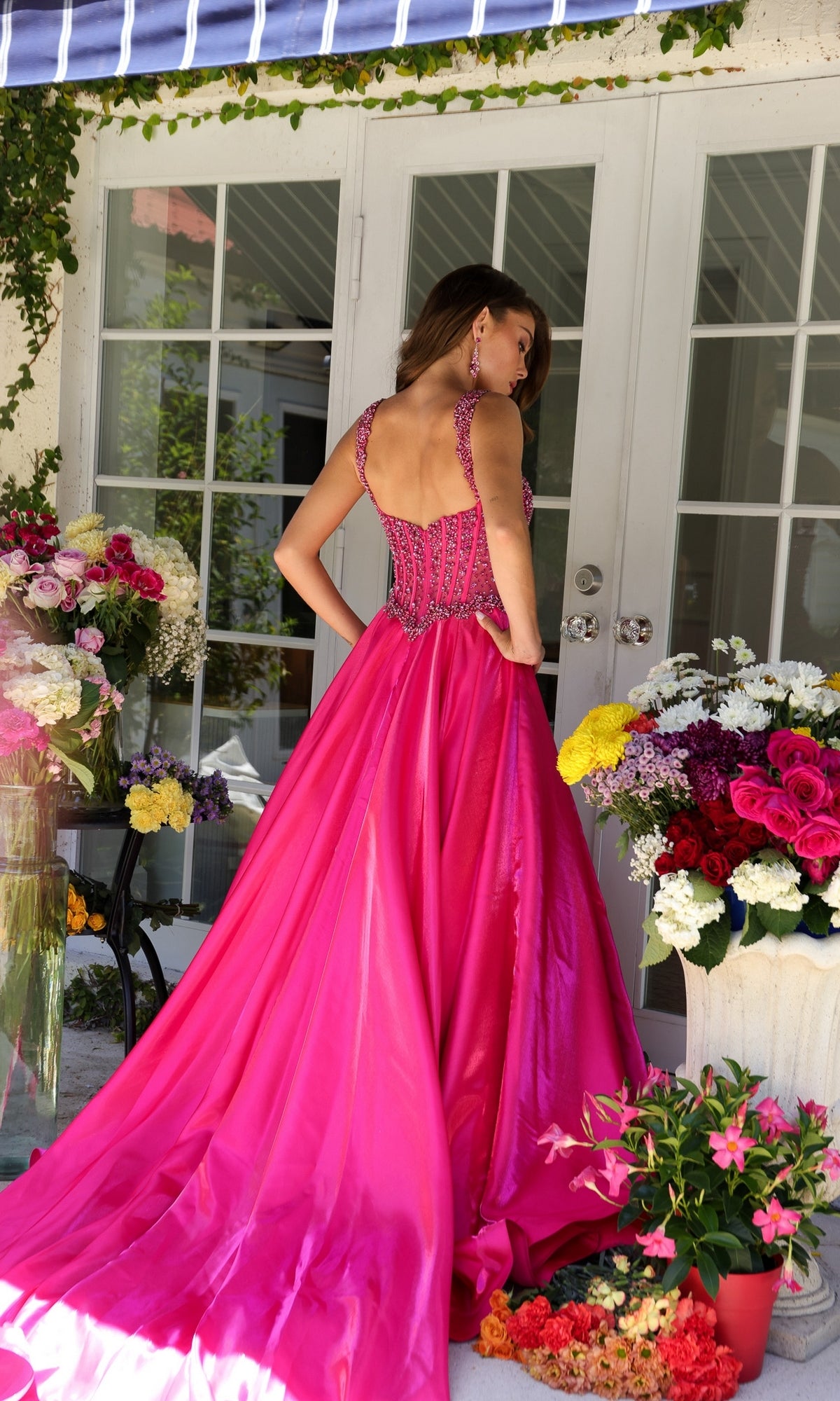 A woman in the Long Formal Dress 29521 by Ava Presley, featuring a bright pink beaded corset bodice and floor-length skirt, stands with her back turned amid vibrant flower arrangements on a patio before glass doors.